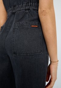 Svart denimromper med elastisk midje, synlige baklommer og rød "Santa Cruz" logo-lapp. Myk tekstur, tettsittende design med avsmalnende form.