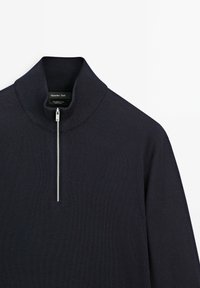 Donkerblauwe pullover met een hoge kraag en een kwart ritssluiting. Gemaakt van merinowol, met een soepele, gestructureerde brei. Merklabel zichtbaar.