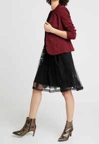 Burgundy sovitusbleiseri mustan pitsireunuksen tyllimekon päällä, yhdistettynä metallisiin nilkkabootseihin, jotka esittelevät kerroksellista, teksturoitua muotoilua.