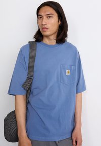 Lyseblå bomulds T-shirt med korte ærmer og en brystlomme. Har et Carhartt logo patch. Gråternet crossbody taske hængende over skulderen.
