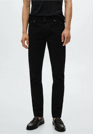 Mężczyzna ubrany w czarne, dopasowane jeansy, czarne loafersy i włożoną do środka czarną koszulę, stojący na tle jasnego, jednolitego tła.