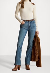 Žena v krémovém roláku, modrých džínách, hnědém pásku a botách, která v jedné ruce drží hnědou koženou bundu.