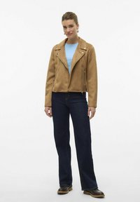 Veste de moto en daim beige avec des fermetures éclair en argent, design ajusté et grands revers, associée à un haut bleu clair et un jean foncé.