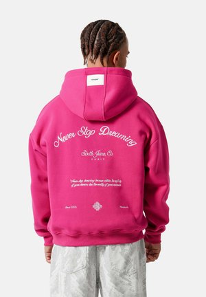 Persoon met gevlochten haar die een roze capuchontrui draagt met witte tekst "Never Stop Dreaming" op de achterkant, gecombineerd met lichtgrijze broek met patroon.