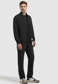 Schwarze Bomberjacke mit gerippten Bündchen und einem Frontreißverschluss, über einem schwarzen Shirt getragen; kombiniert mit dunklen Jeans und mehrfarbigen Sneakers.