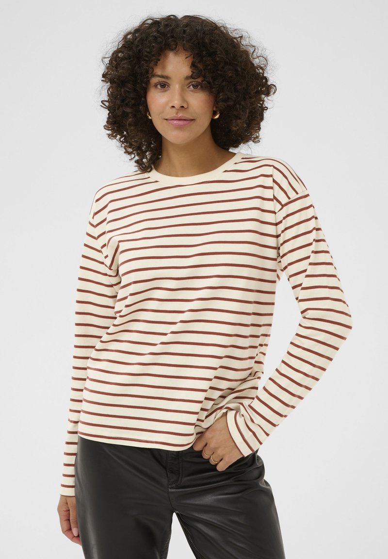 Vrouw met krullend haar, gekleed in een crèmekleurig shirt met lange mouwen met bruine horizontale strepen en zwarte leren broek, staand met één hand in haar zak.