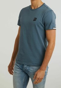 Muž v modrém tričku s krátkým rukávem a kulatým výstřihem, na kterém je malá černá logo na hrudi a pruhovaná detaile na rukávu, v kombinaci se světle modrými džíny.