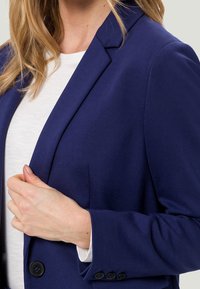Blazer bleu marine à la coupe structurée, fermeture simple boutonnage, et boutons noirs. Confectionné en tissu lisse avec une coupe cintrée.