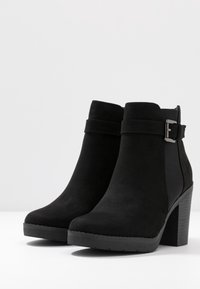 Botines negros con una textura similar al ante, un tacón cuadrado y una correa decorativa con una hebilla de plata en el lado exterior.