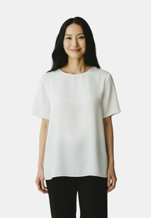 Donna sorridente con lunghi capelli neri che indossa una camicetta bianca a maniche corte ampia e pantaloni neri su uno sfondo neutro.