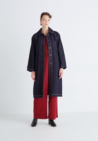 Dunkelblauer Denim-Langmantel mit weitem Kragen, Druckknopfverschluss, kontrastierenden gelben Nähten, getragen über einem roten Outfit und schwarzen Stiefeln.