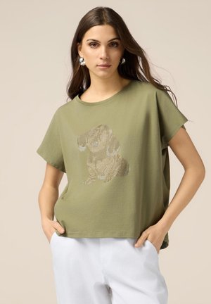 Camiseta de algodón verde oliva de manga corta con un gráfico de perro brillante en tonos metálicos, combinada con pantalones blancos.