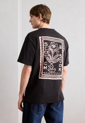 Ung mand iført sort t-shirt med pink blomstergrafik og teksten "Scotch & Soda Amsterdam" på ryggen, kombineret med blå jeans.
