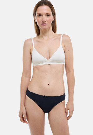 Femme debout, de face, portant une bralette blanche et une culotte noire sur un fond uni clair.