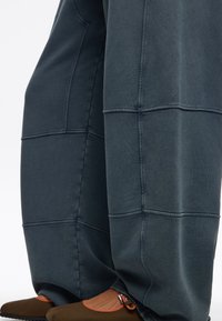 Pantalones de pierna ancha en verde azulado oscuro, hechos de una tela suave con detalles de paneles cosidos. Combinados con zapatos marrones que presentan un acento naranja.