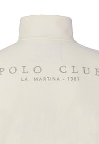Camisa de cuello alto color crema con texto bordado "POLO CLUB LA MARTINA - 1987" en la parte trasera cerca del cuello.