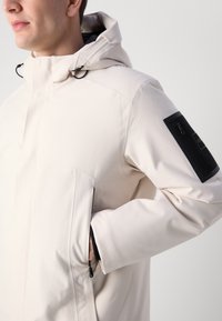 Lys beige jakke med en tekstureret overflade, der har hætte, sidelommer og en sort lynlåslomme på ærmet.