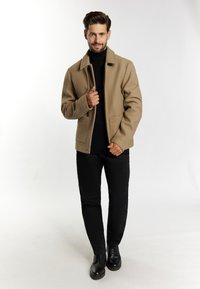 Jachetă din material lână cu tonuri de bej, cu guler larg, nasturi în față și două buzunare, purtată cu un pulover negru cu guler înalt și pantaloni negri, completată cu pantofi negri.