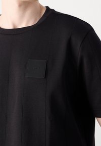 Chemise noire à manches courtes avec des détails de coutures verticales et un petit patch carré texturé sur la poitrine gauche.