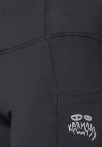 Juodos sportinės kelnės, turinčios lygią tekstūrą, su kontrastinga siuvimo linija ir balta spausdinta logotipu. Matoma šoninė kišenė.
