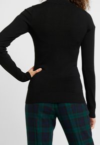 Musta pitkähihainen neulepaita korkealla kauluksella, istuvalla mallilla ja resoreilla hihoissa ja helmassa. Yhdistetty vihreisiin ja sinisiin ruudullisiin housuihin.