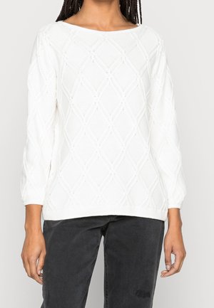 Kvinde iført en hvid tekstureret langærmet sweater med diamantmønstre og løse sorte bukser, vist fra skuldre til hofter.