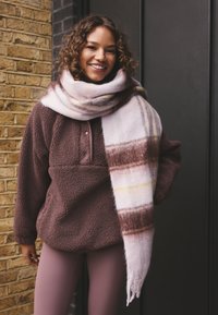 Brun fleecetröja med knäppning vid halsen, kombinerad med en rosa och brunrandig halsduk samt ljusrosa leggings mot en tegelvägg.