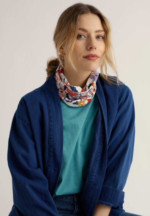 Joven con cabello rubio que lleva un pañuelo floral en el cuello, camisa verde azulado y chaqueta holgada azul oscuro sobre un fondo claro y liso.