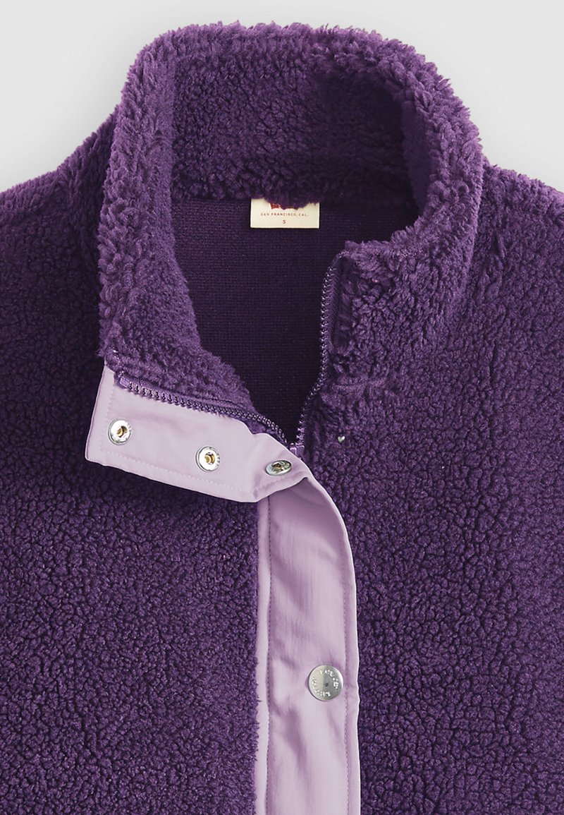 Veste polaire violette avec un col montant, fermeture à pression et une doublure intérieure lilas contrastante. Texture douce avec coutures visibles et détails métalliques.