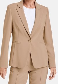 Blazer beige ajusté avec un seul bouton, revers crantés et deux poches avant. Porté sur un haut blanc côtelé, texture de tissu lisse.