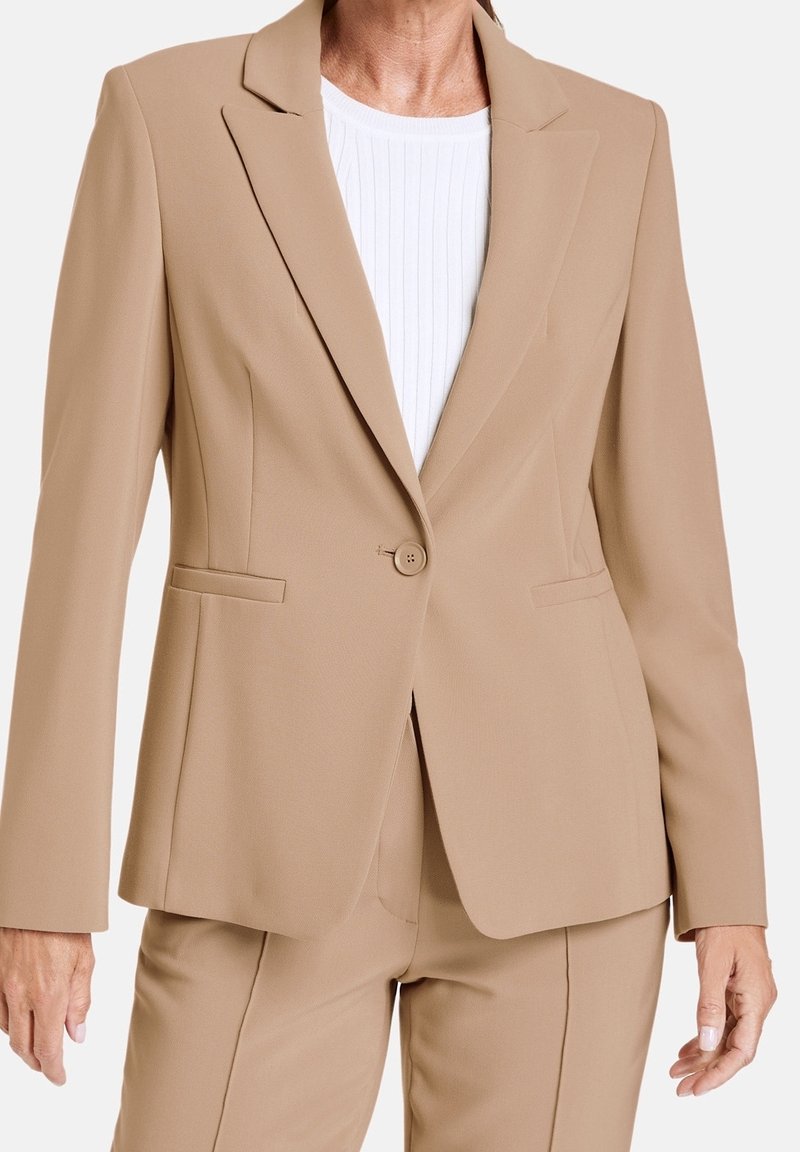 Blazer beige ajusté avec un seul bouton, revers crantés et deux poches avant. Porté sur un haut blanc côtelé, texture de tissu lisse.