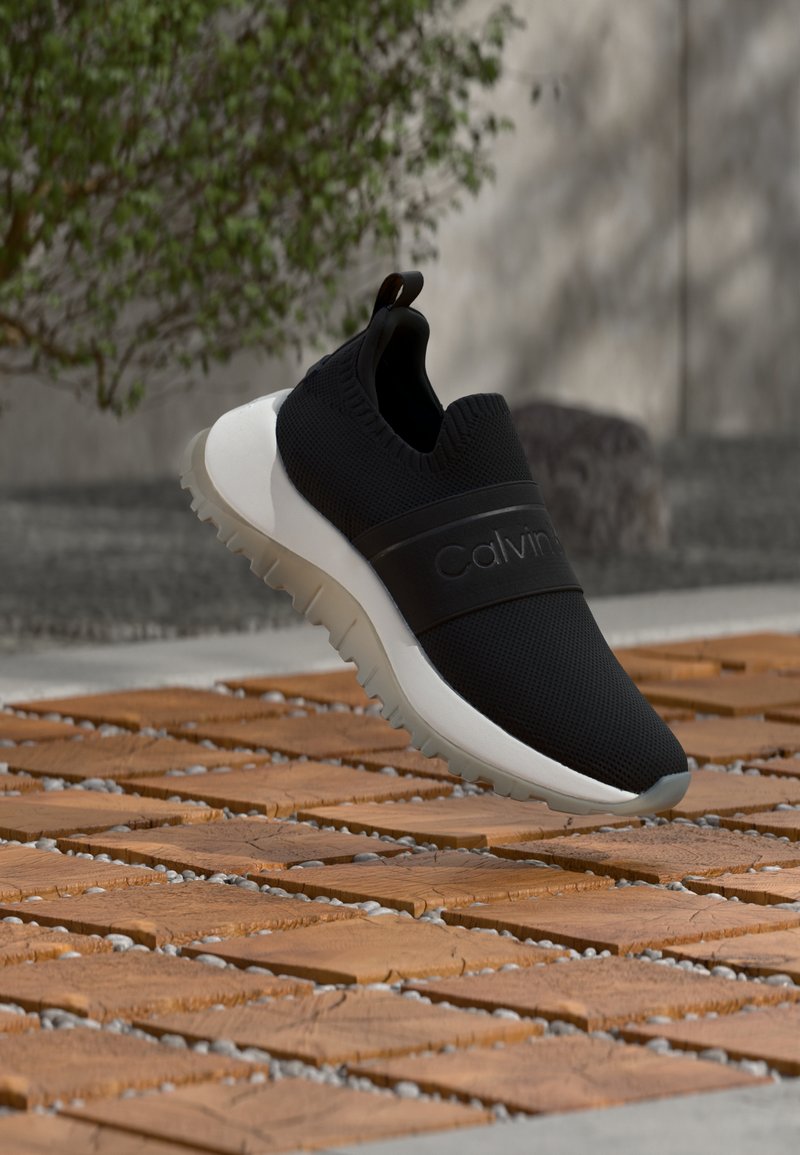 Chaussure de sport noire sans lacets avec semelle blanche et grip texturé, flottant au-dessus d'une surface en carreaux de bois en plein air près de la verdure.