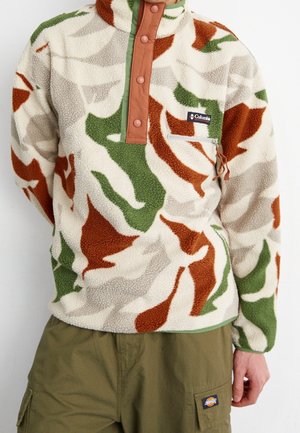 Person iført en fleece-pullover med grønne, brune og beige bladmønstre, en skjult knapoplukning og olivengrønne cargobukser.