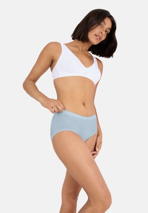 Vrouw in wit draadloos beha en lichtblauwe katoenen slip, poseert tegen een effen lichte achtergrond.