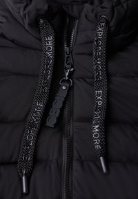 Svart pufferjacka med kviltad textur, utrustad med flätade dragsnören med texten "UTFORSKA MER" och en svart dragkedja med en märkeslogga på dragfliken.