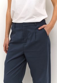Pantalon marine à coupe ajustée, avec des plis sur le devant, des poches sur les côtés et un tissu lisse et texturé. Porté avec un haut blanc.