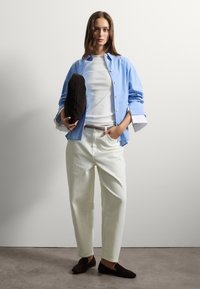 Joven mujer de pie contra una pared blanca, vistiendo una camisa de color azul claro, una blusa blanca, pantalones blancos, mocasines negros y sosteniendo un bolso de mano negro.
