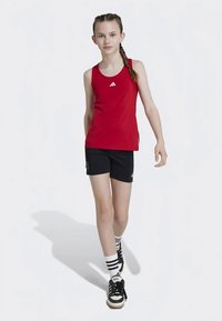 Rødt atletisk singlet med hvit logo, svarte shorts og svarte joggesko med hvite striper; modellen har fletninger og står selvsikkert.