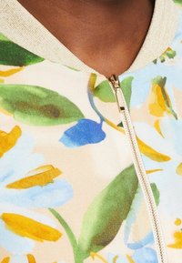 Blomstret lynlåsjakke i blød kvalitet med grønne, blå og gule mønstre. Den har en lynlås i gyldent farve og en struktureret, lysfarvet krave.