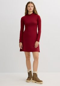 Vestido rojo de manga larga con un corsé ajustado, dobladillo acampanado y tejido suave. Llevado con botas de piel sintética de color marrón claro con forro, que presentan cordones y una suela gruesa.