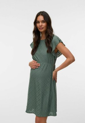 Gravid kvinde med langt mørkt hår iført en grøn kjole med hulmønster, der blidt holder sin mave og står mod en ensfarvet baggrund.