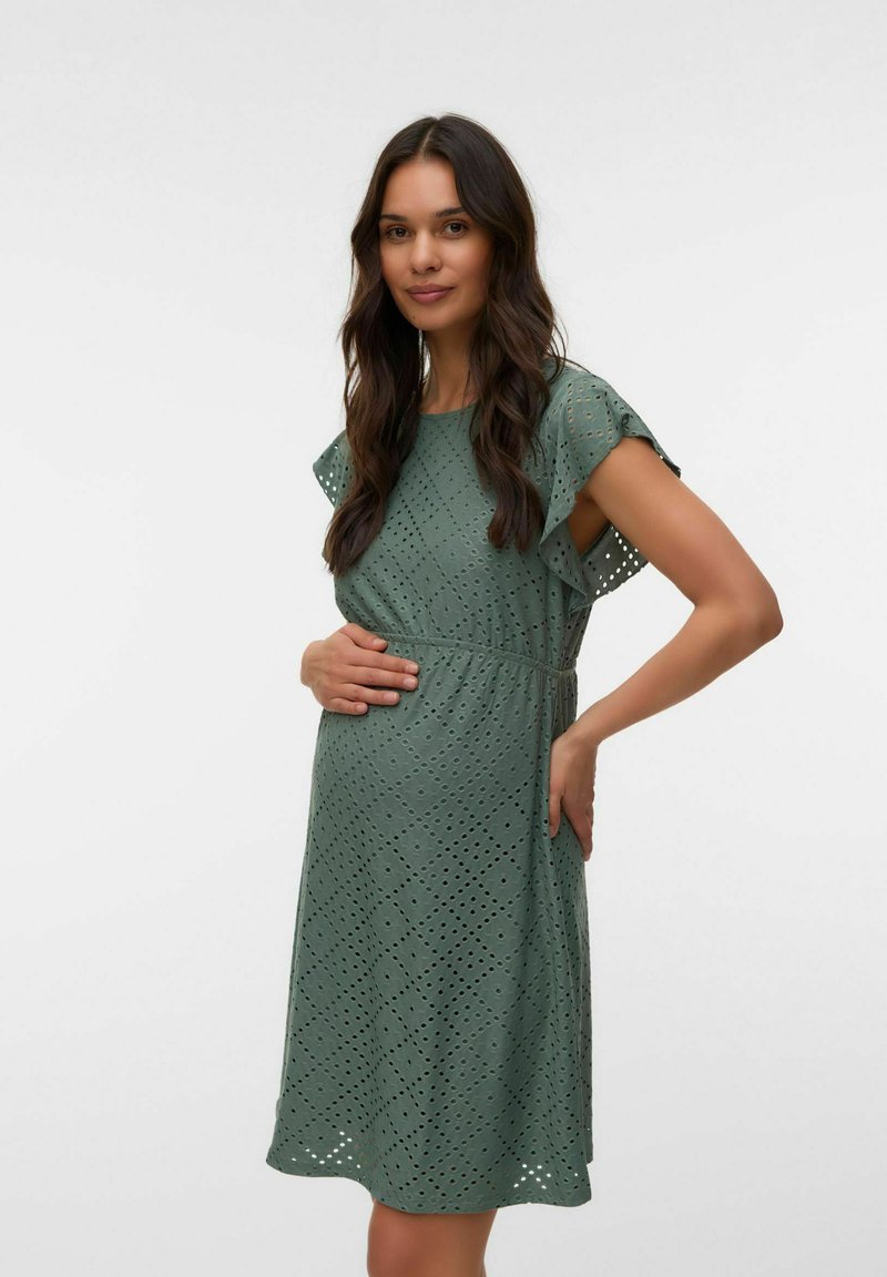 Femme enceinte aux longs cheveux foncés portant une robe verte à motif œillet, tenant doucement son ventre et posant devant un fond uni.