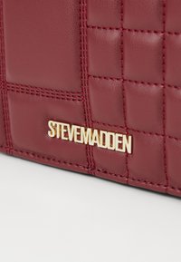 Lähikuva viininpunaisesta tikkikuvioidusta nahasta, jossa kultainen "Steve Madden" -logo käsilaukun kulmassa.