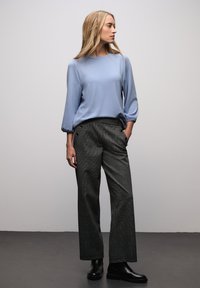 Jasnoniebieska bluzka z długim rękawem ze ściągaczami przy mankietach, zestawiona z wysokim stanem spodni w kratę z bocznymi guzikami oraz czarnymi butami.