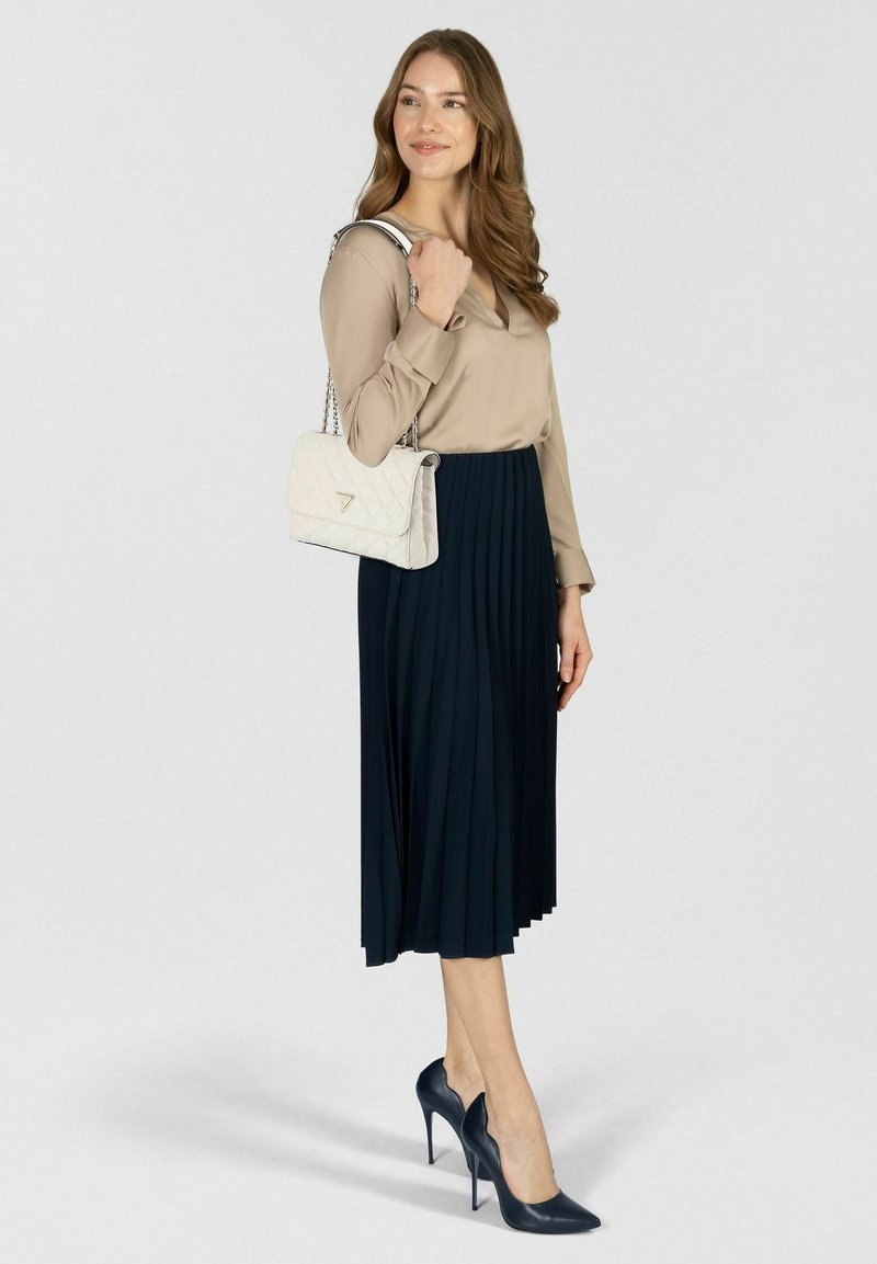 Beige blouse met lange mouwen, gecombineerd met een geplooide marineblauwe rok. Accessoires zijn een witte doorgestikte handtas en donkere hoge hakken.