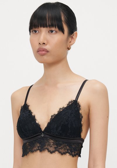 Zwarte kant bralette met driehoekige cups, dunne bandjes, een geschulpte kantafwerking en een stoffen band onder de buste. Gelaagd kantpatroon overal.