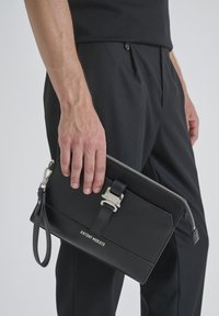 Clutch de cuero negro con herrajes plateados, que presenta una cremallera en la parte superior, una correa con detalle de hebilla y el nombre de la marca en relieve en la parte frontal.