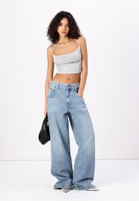 Top din plasă gri, cu un decolteu rotund, asortat cu blugi largi, de culoare albastră deschis. Modelul ține o geantă neagră. Blugii au talie înaltă și un ușor efect de uzură.