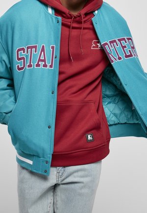 Person trägt eine türkisfarbene Varsityjacke über einem weinroten Hoodie und hellblauen Jeans, die Jacke ist teilweise geöffnet und zeigt ein gestepptes Futter.