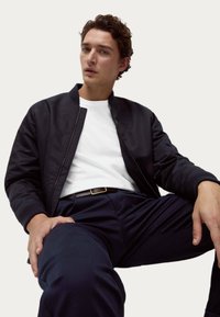 Černá bomber bunda s žebrovanými manžetami, bílý tričko, námořnické kalhoty a hnědý opasek. Model sedí na světlém pozadí.